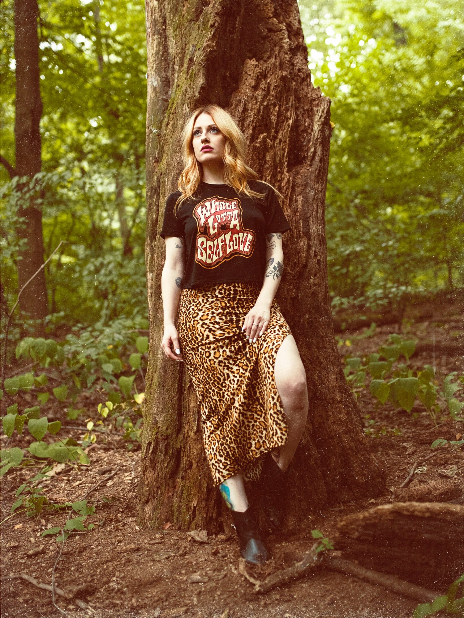 A female model wearing a black crop top with the graphic 'Whole Lotta Self Love' standing next to a tree.