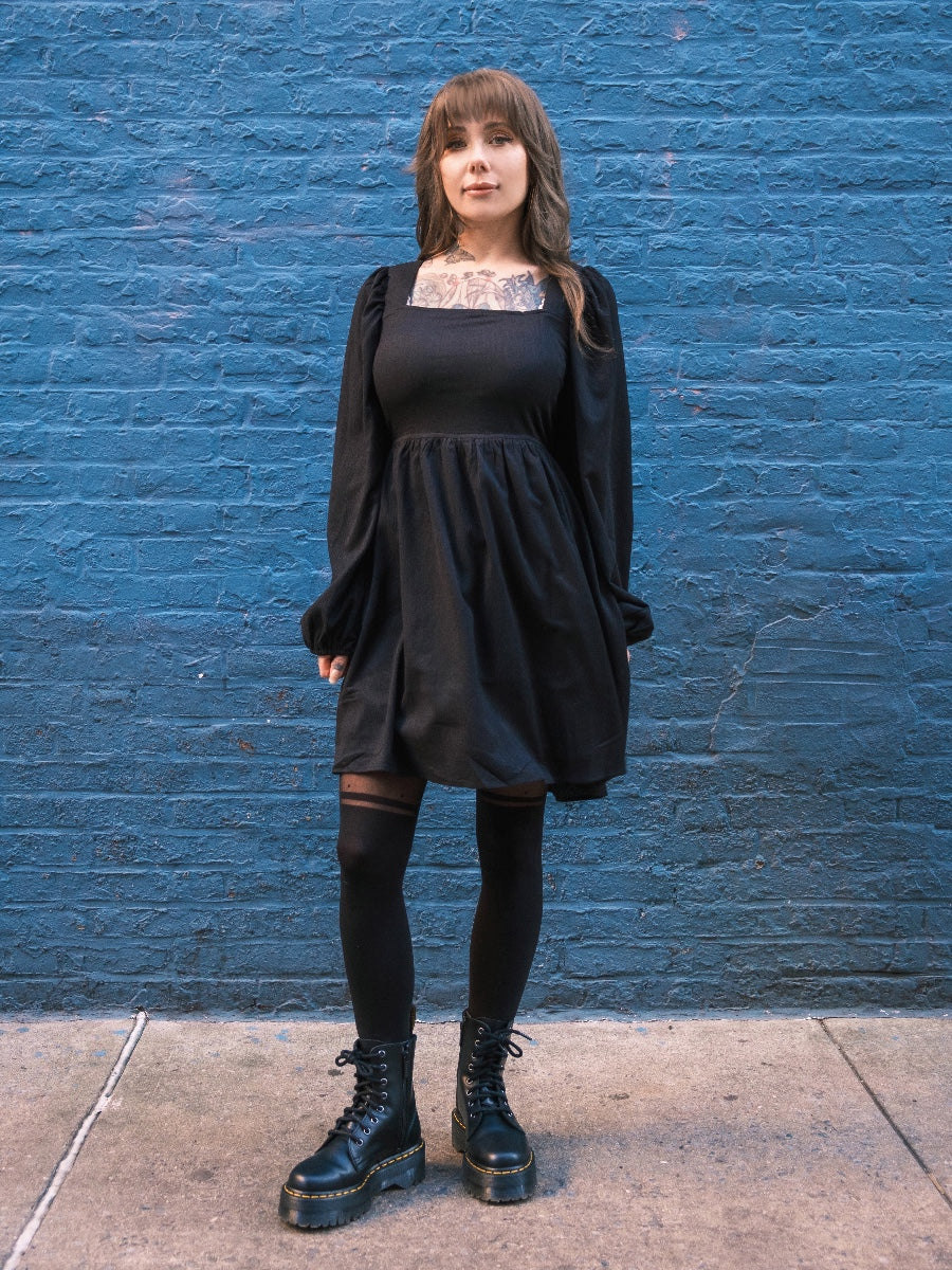 Megan Massacre wearing a black longsleeve dress against a blue brick wall