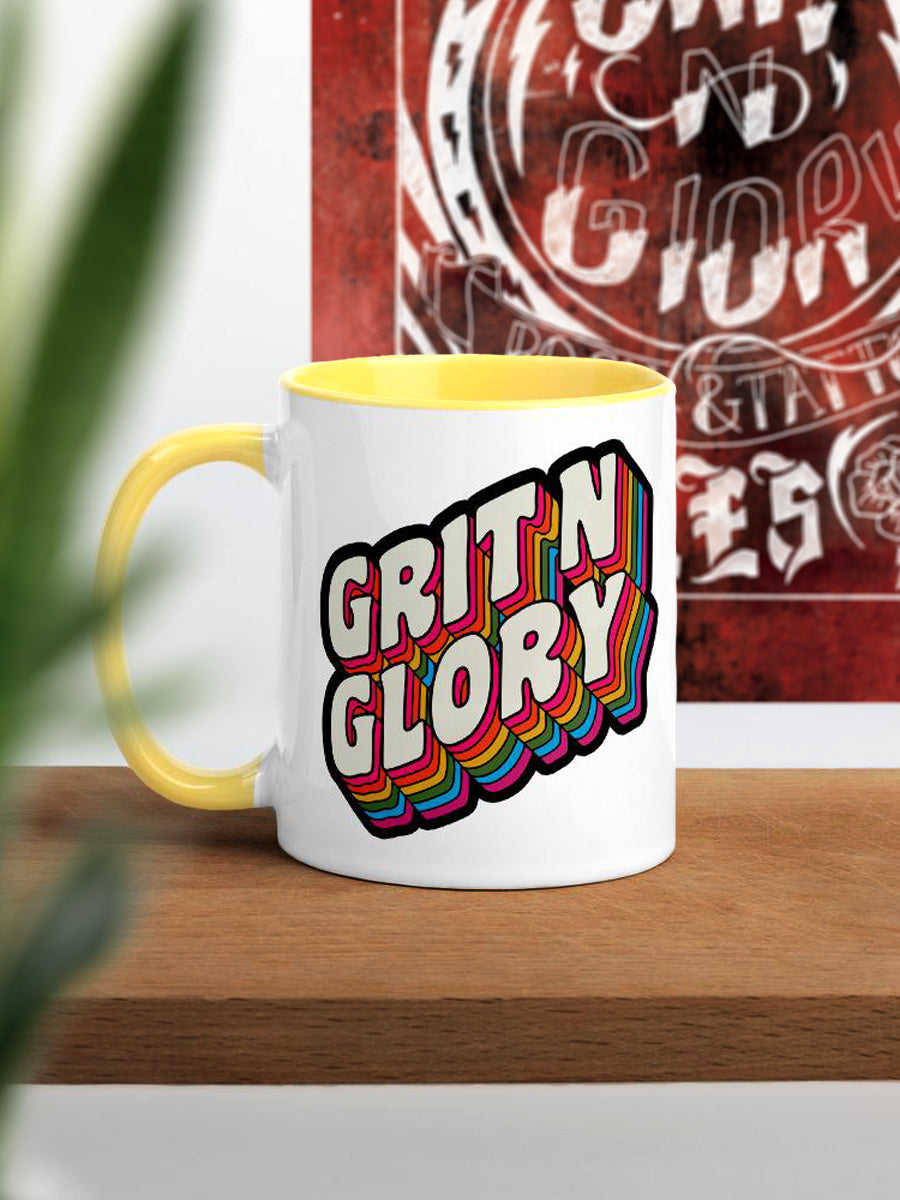A white and yellow ceramic mug with a colorful 'Grit N Glory' logo print.